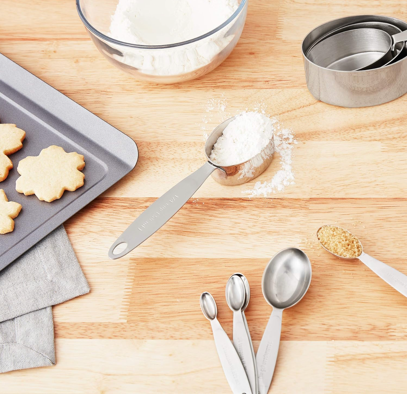 Baking utensils including a tray of cookies, measuring cups, and spoons on a wooden surface.